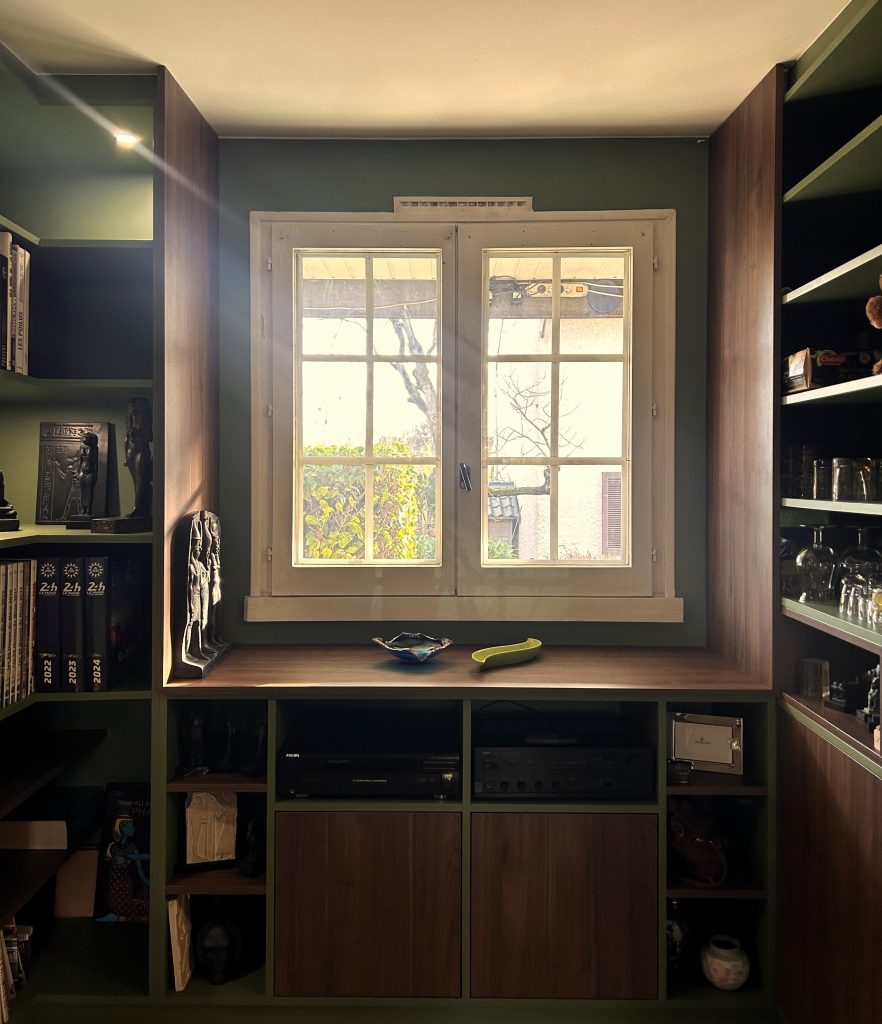 Bibliothèque sur-mesure vert olive encadrant une fenêtre, avec rangement en noyer et objets décoratifs.