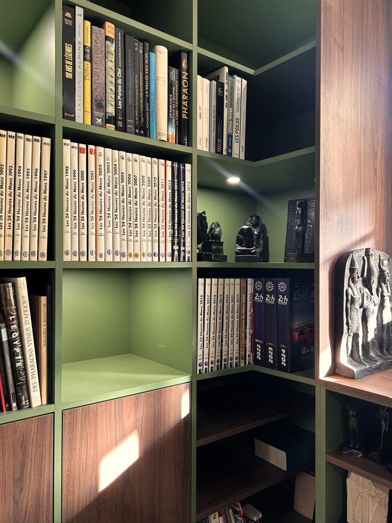 Bibliothèque murale sur-mesure vert olive et noyer entourant une fenêtre centrale, réalisée par Lunello.